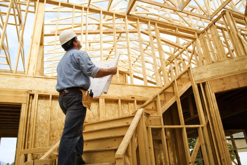 Inspector with hard hat and blueprints reviewing roof trusses and structural framing inside a custom home during a framing inspection