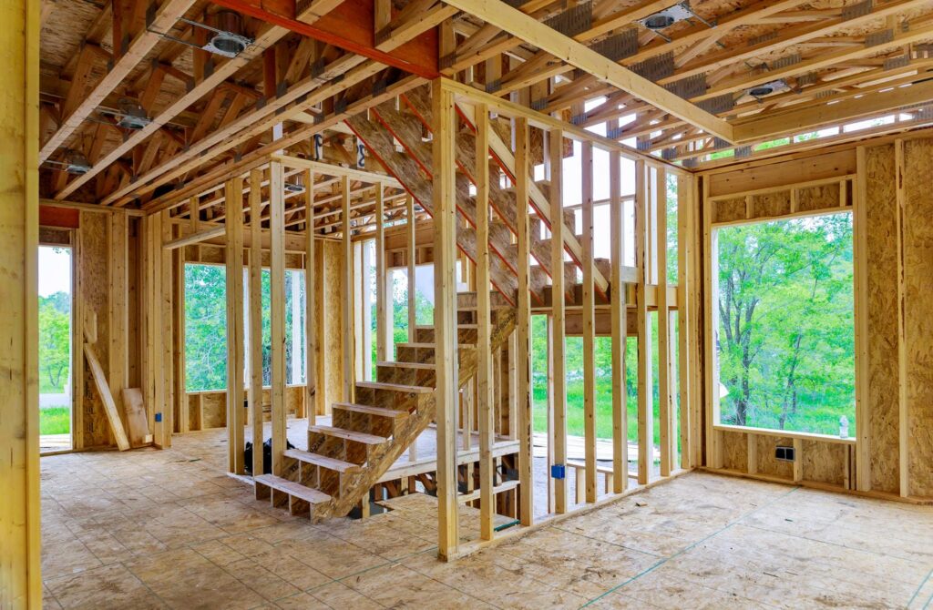 Interior of a custom home in the framing stage showing engineered I-joists, beams, and dimensional lumber wall studs supporting an open floor plan with large window openings and a stair opening