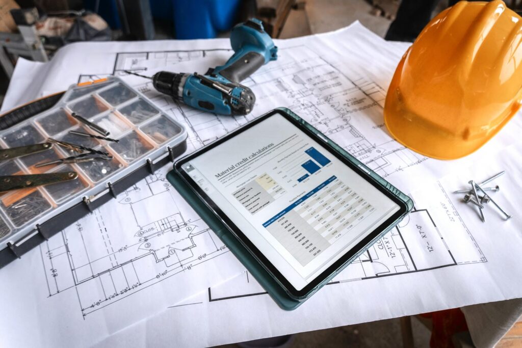 Construction blueprints, tablet displaying material calculations, hard hat, and tools laid out on a jobsite table in preparation for a framing inspection